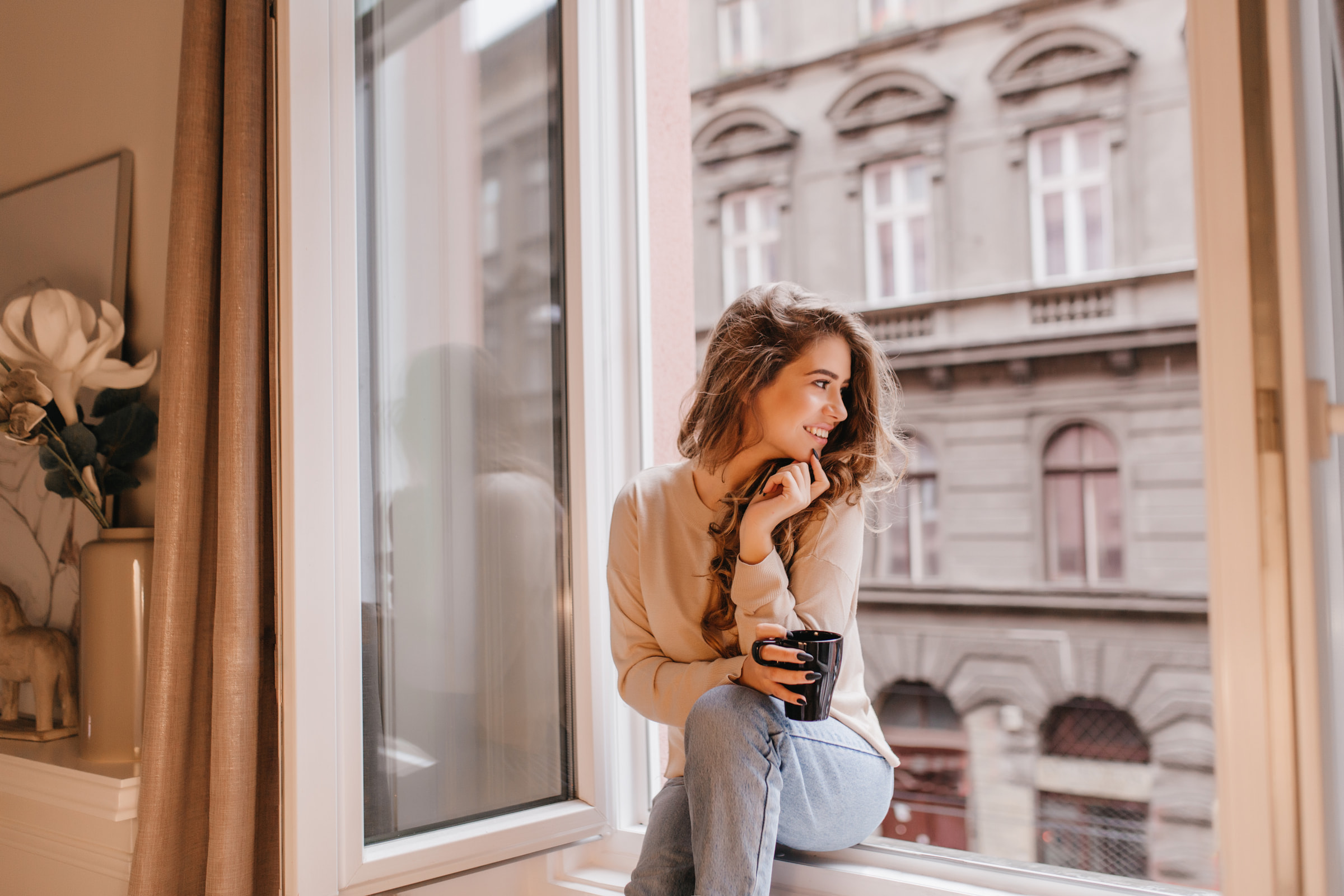 Uśmiechnięta kobieta na parapecie trzyma kubek, za szybą widok miasta