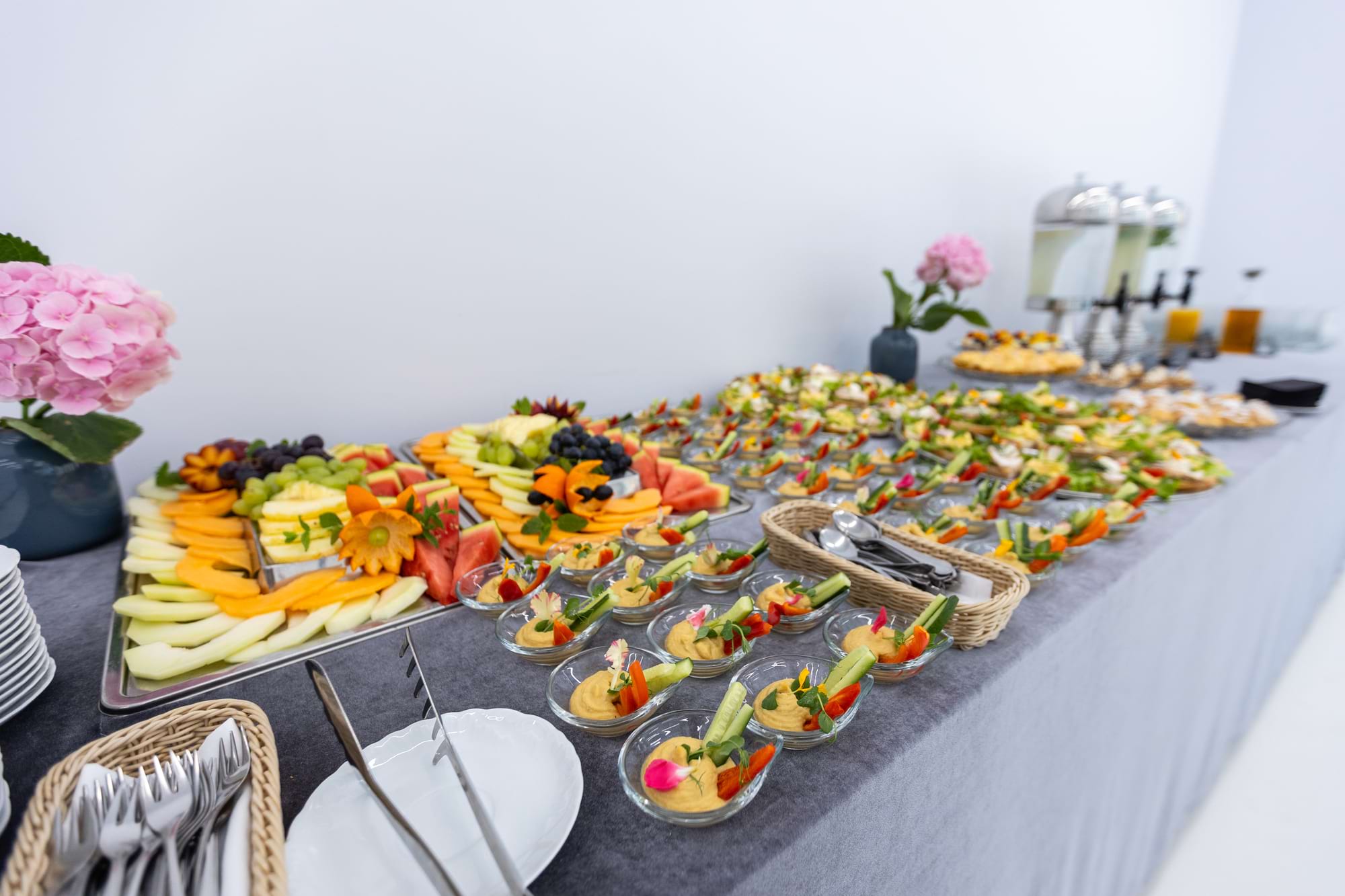 Długi stół z przekąskami i owocami, ozdobiony różowymi hortensjami