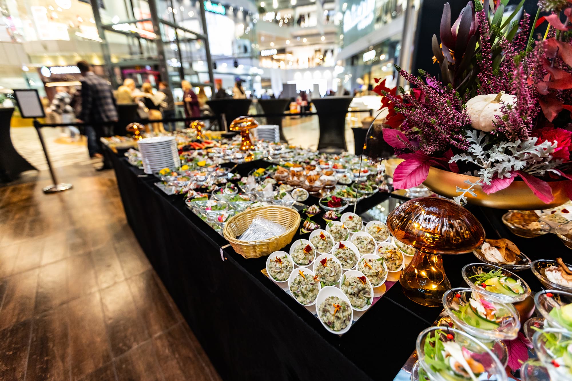 Duży bufet przekąsek z kwiatową dekoracją po prawej w galerii handlowej
