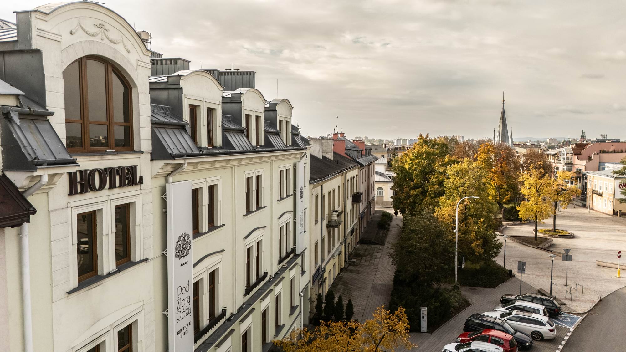 Hotel na lewo, ulica z autami i jesiennymi drzewami, wieża kościoła